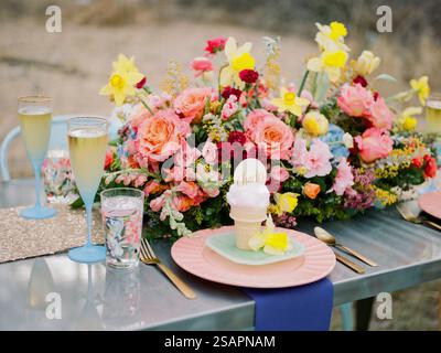 Un tavolo all'aperto splendidamente allestito presenta un vivido pezzo centrale floreale insieme a piatti pastello, bicchieri di bevanda frizzante e un delizioso dessert Foto Stock