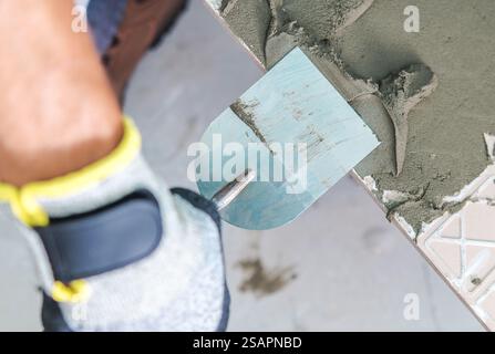 Un lavoratore applica cemento con una cazzuola su una superficie piastrellata in un cantiere durante il giorno, concentrandosi sulla precisione e la qualità. Foto Stock