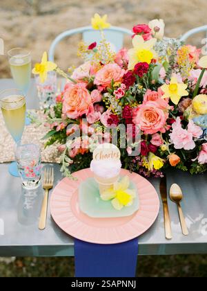 Un tavolo all'aperto splendidamente allestito presenta un piatto pastello con un dessert personalizzato, fiori vivaci ed eleganti bicchieri, perfetto per una celebrità Foto Stock