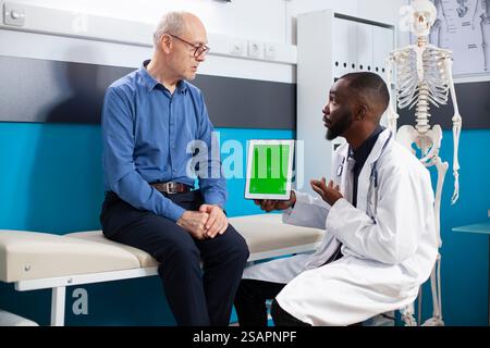 Il paziente anziano ascolta mentre il medico spiega il piano di trattamento che tiene in mano una compressa con schermo verde. Il medico consulta il vecchio sulla sua condizione medica in clinica utilizzando dispositivo con display mockup isolato. Foto Stock