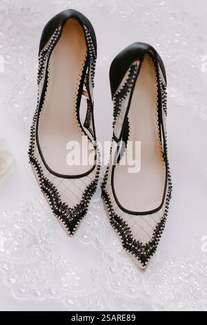 Un paio di eleganti tacchi alti bianchi e neri sono esposti su un delicato sfondo in pizzo. Le scarpe sono caratterizzate da perline intricate e punta a punta, Foto Stock