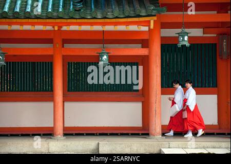 La fanciulla del Santuario ('miko') vestita con tradizionale hakama rosso e kosode bianco che cammina sotto un corridoio coperto al santuario shintoista Heian Jingu, Kyoto, Giappone. Foto Stock