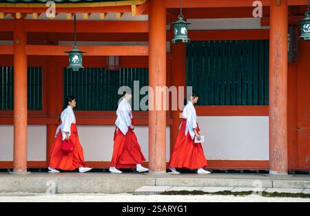 La fanciulla del Santuario ('miko') vestita con tradizionale hakama rosso e kosode bianco che cammina sotto un corridoio coperto al santuario shintoista Heian Jingu, Kyoto, Giappone. Foto Stock