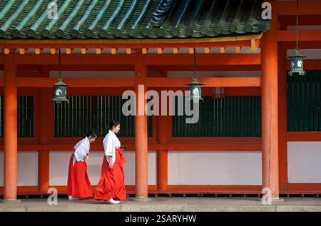 La fanciulla del Santuario ('miko') vestita con tradizionale hakama rosso e kosode bianco che cammina sotto un corridoio coperto al santuario shintoista Heian Jingu, Kyoto, Giappone. Foto Stock