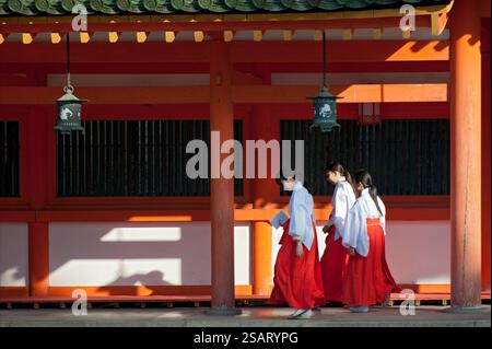 La fanciulla del Santuario ('miko') vestita con tradizionale hakama rosso e kosode bianco che cammina sotto un corridoio coperto al santuario shintoista Heian Jingu, Kyoto, Giappone. Foto Stock