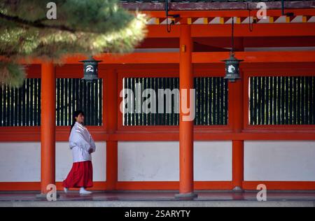 La fanciulla del Santuario ('miko') vestita con tradizionale hakama rosso e kosode bianco che cammina sotto un corridoio coperto al santuario shintoista Heian Jingu, Kyoto, Giappone. Foto Stock