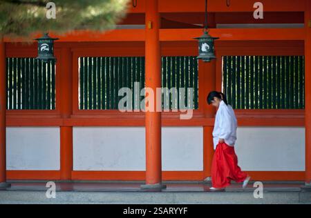 La fanciulla del Santuario ('miko') vestita con tradizionale hakama rosso e kosode bianco che cammina sotto un corridoio coperto al santuario shintoista Heian Jingu, Kyoto, Giappone. Foto Stock