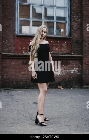 Donna con un vestito nero che posa contro un muro rustico di mattoni Foto Stock