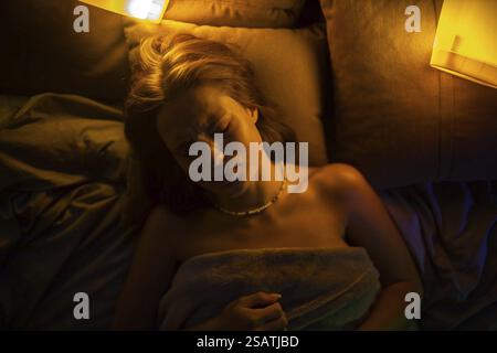 Una donna in difficoltà giace su cuscini marroni nel letto, illuminata da due luci con bagliori contrastanti Foto Stock
