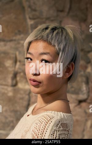 Giovane donna che mostra un taglio di capelli argentati corti alla moda davanti a un muro di pietra Foto Stock