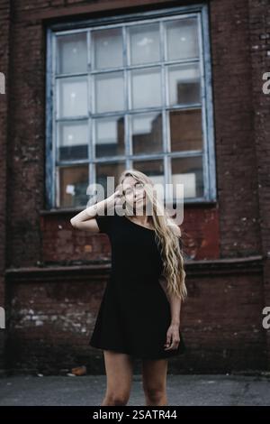 Una donna con un abito nero posa graziosamente di fronte a un muro urbano di mattoni Foto Stock