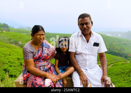 Una famiglia di tre persone è seduta insieme su una panchina di pietra in mezzo a un lussureggiante paesaggio verde. La donna indossa un sari colorito Foto Stock