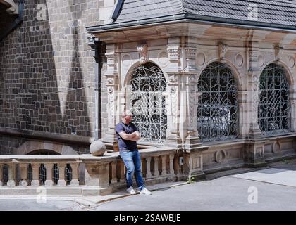 Uomo con occhiali, mani piegate sul petto in piedi davanti al castello, vestito di jeans, sneakers bianche e una polo blu, un castello medievale fatto di Foto Stock