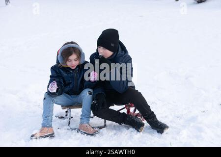 Un ragazzo e una ragazza, vestiti caldi con giacche e cappelli invernali, siedono su una piccola slitta in un paesaggio innevato. Sembra che stiano godendo di una tranquilla giornata invernale Foto Stock