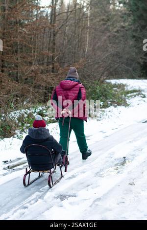 Una persona con una giacca rossa tira un bambino su una piccola slitta lungo un sentiero innevato. Il sentiero si snoda attraverso una foresta invernale senza fronzoli. La scena è pacifica Foto Stock