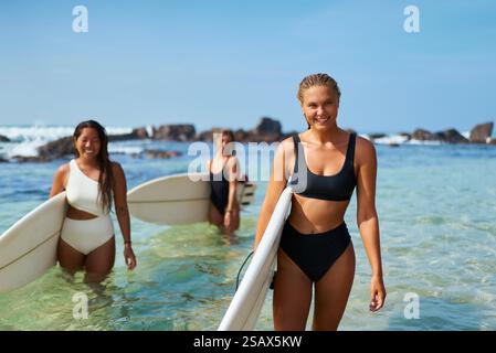 Tre donne in costume da bagno tengono le tavole da surf nelle soleggiate acque dell'oceano. Gli amici amano le calde avventure in riva al mare, fare surf insieme su una spiaggia esotica. Vivace e divertente Foto Stock