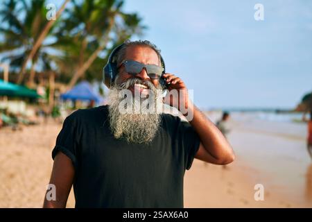L'uomo con la barba grigia, occhiali da sole, ascolta la musica con le cuffie sulla spiaggia soleggiata. Il nonno elegante ama le melodie, sentirsi giovane e vibrante Foto Stock