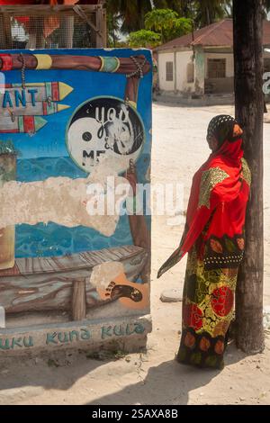 Africa Tanzania, Zanzibar, costa orientale, Jambiani, villaggio, donna locale vestita in colori, accanto al murale del ristorante Foto Stock