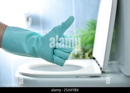 Bagno appena disinfettato con mano con guanti in gomma blu che danno il pollice verso l'alto gesto e ciotola WC sullo sfondo con coperchio aperto blu in piastrelle b Foto Stock