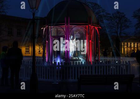 Statua illuminata in un padiglione di vetro di notte, circondata da luci colorate. I visitatori sagomati ammirano le opere d'arte di Harrogate, Regno Unito. Foto Stock