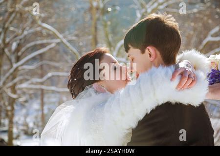 Sposa e sposo abbracciano tenermente in un ambiente innevato all'aperto, circondato da alberi la sposa indossa la stola di pelliccia, Foto Stock