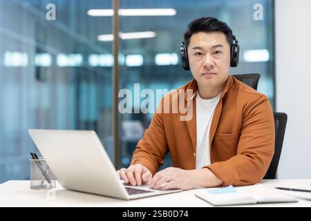 Ritratto di un serio programmatore asiatico maturo. Uomo concentrato con cuffie e laptop che guarda la fotocamera, sviluppatore sul posto di lavoro all'interno dell'ufficio aziendale vicino alla finestra. Foto Stock