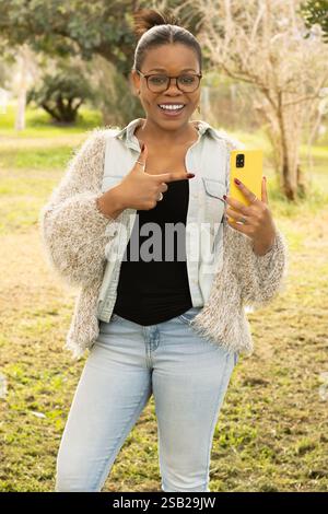 Una donna nera allegra con occhiali e un outfit elegante è in piedi all'aperto, tenendo in mano uno smartphone giallo e puntandolo con entusiasmo. Foto Stock