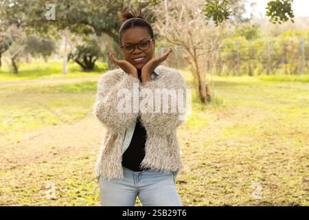 Una donna nera gioiosa con gli occhiali posa giocosa con le mani sotto il mento mentre sorride. Foto Stock
