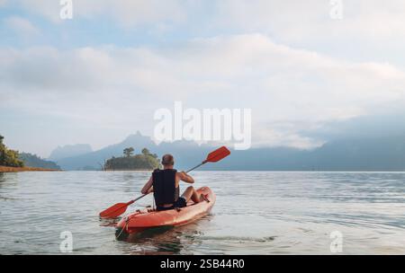 Uomo che pagaia in kayak sul calmo lago Cheow LAN e incontra l'alba nel parco nazionale di Khao Sok, Thailandia. Acque tranquille, luce dorata e scogliere calcaree creano Foto Stock