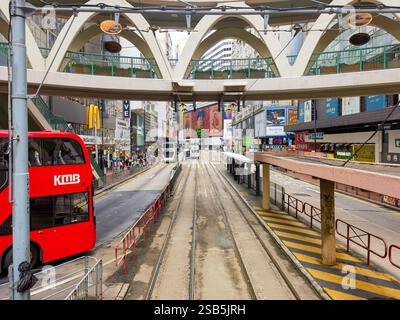 Hong Kong, Cina - 1 febbraio 2025: Un autobus rosso a due piani si trova accanto ai binari del tram di Hong Kong sotto un moderno cavalcavia. Edifici cittadini e negozi li Foto Stock