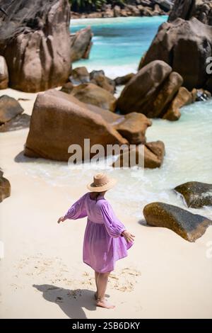 Una donna con un abito rosa e un cappello di paglia cammina lungo una spiaggia sabbiosa vicino a grandi rocce di granito, con acqua turchese sullo sfondo Foto Stock
