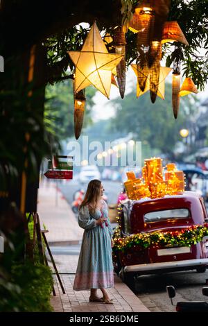 Una donna si erge su una strada decorata con lanterne scintillanti delle stelle natalizie appese sopra e un'auto d'epoca adornata con scatole regalo e luci festive Foto Stock
