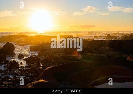 Un caldo e dorato tramonto illumina la costa rocciosa di una spiaggia sudafricana. Le onde si infrangono dolcemente mentre le sagome di due figure si trovano sulle rocce Foto Stock