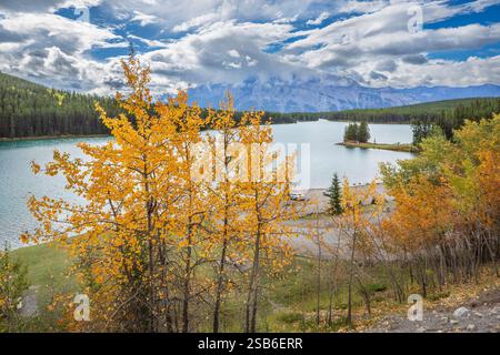 Two Jack Lake in Canada, circondato da vibranti alberi autunnali, calme acque turchesi e maestose montagne sotto un cielo nuvoloso dinamico. Foto Stock