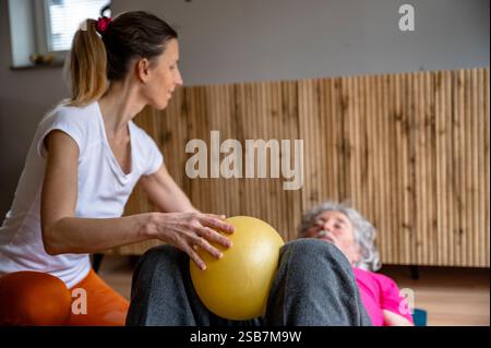 Un terapeuta assiste una persona anziana con un esercizio riabilitativo utilizzando una palla gialla per il rafforzamento delle gambe. Foto Stock