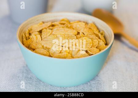 Granoturco croccante non dolcificato in ciotola blu, latte, tazza e cucchiaio sul retro, fotografati con luce naturale (messa a fuoco selettiva) Foto Stock