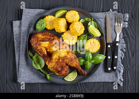 metà di pollo arrosto con pannocchie di mais monete e foglie di basilico fresco sul piatto su un tavolo di legno nero con forchetta e coltello, vista orizzontale dall'alto, fl Foto Stock