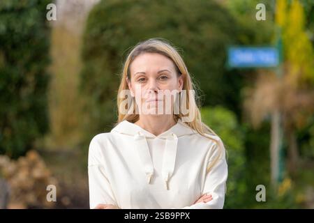 Una donna con i capelli biondi sta all'aperto con le braccia incrociate, guardando direttamente la fotocamera con un'espressione seria e mirata. Foto Stock