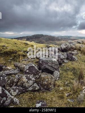 Le pietre ricoperte di muschio formano un confine naturale su una collina che domina un terreno ondulato sotto un cielo nuvoloso. Il paesaggio trasmette un tranquillo voi Foto Stock