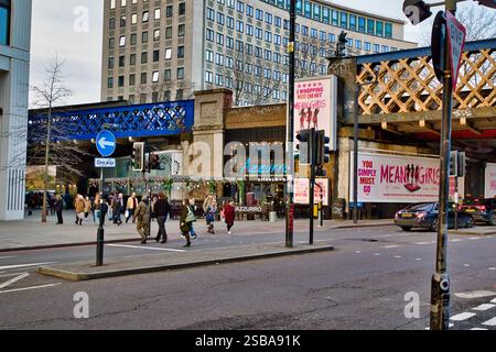 Una scena di strada cittadina con pedoni che attraversano un incrocio, vari cartelli stradali, edifici, un ponte di metallo blu e cartelloni pubblicitari Foto Stock