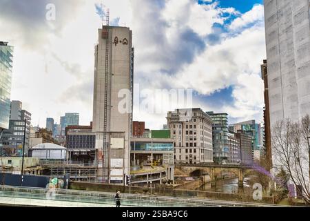 Paesaggio urbano caratterizzato da un alto edificio grigio chiaro in costruzione, adiacente a vecchi edifici in mattoni e un corso d'acqua, sotto un cielo parzialmente nuvoloso. Foto Stock