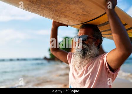 Un anziano uomo etnico con tavola da surf si trova sulla spiaggia sabbiosa. I surfisti maschi maturi amano il sole e la brezza dell'oceano mentre si preparano per le sessioni di surf. Attivo Foto Stock