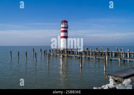 Un faro a righe rosse e bianche si erge a sentinella su un molo di legno che si estende in un tranquillo lago. Cielo soleggiato e acqua placida. Foto Stock