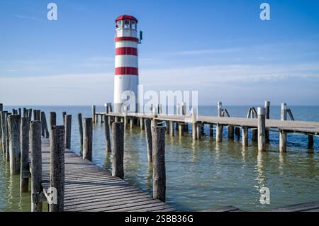 Il molo in legno si estende in acque calme, un faro a strisce si erge in lontananza in una giornata di sole. Foto Stock