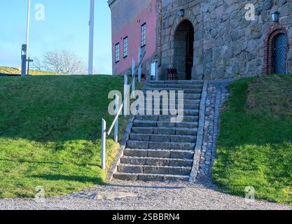 Scalini di pietra intemprati salgono verso un vecchio edificio in pietra grezza, circondato da lussureggianti prati verdi e un cielo blu, a indicare una giornata tranquilla Foto Stock