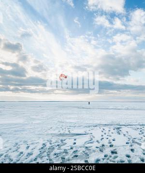 Kiteboarding su un enorme lago ghiacciato coperto di neve fitta. Aquilone rosso che vola nel vento, nuvole sopra l'infinita superficie del deserto di neve con impronte. Foto Stock