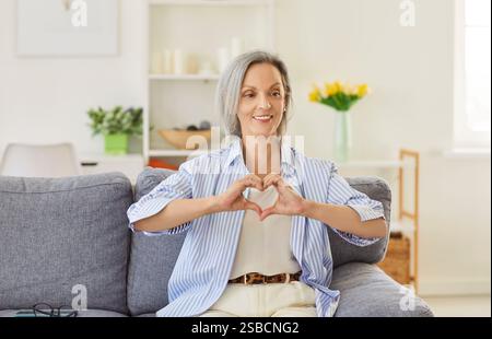 Donna anziana seduta sul divano nel soggiorno di casa e mostra un segno d'amore con le mani che fanno il cuore Foto Stock