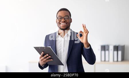 Ritratto di allegro psicologo maschio nero che mostra un buon gesto, raccomandando aiuto professionale in clinica Foto Stock