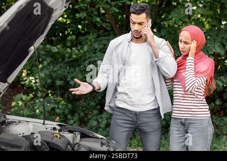 Triste despaired donna araba in hijab e l'uomo chiamata al supporto auto, stare vicino a macchina rotta con cofano aperto Foto Stock
