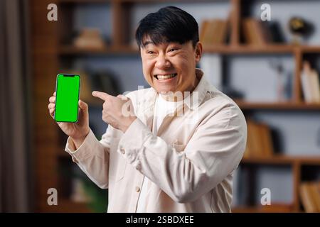 Felice uomo asiatico, mostra dispositivo, offerta online, spazio libero vuoto, uomo allegramente che mostra uno smartphone con uno schermo vuoto mentre indica un ambiente accogliente Foto Stock